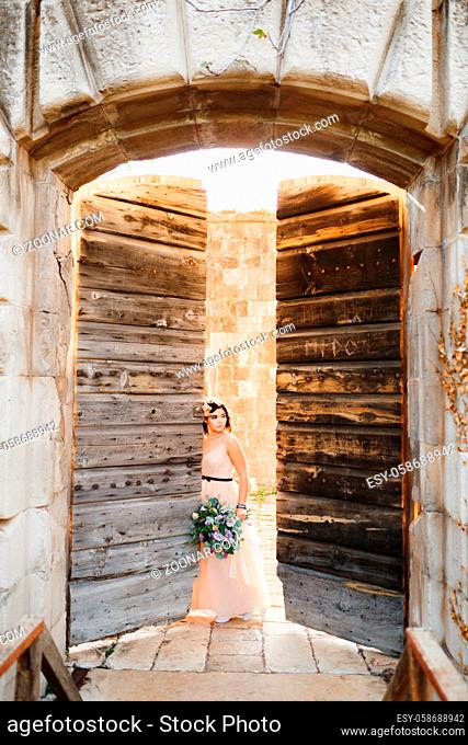 Cute bride in a beautiful dress with a bouquet of flowers is leaning against the open wooden door of the castle. High quality photo