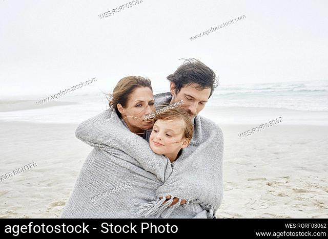 Man and woman standing with daughter on windy beach wrapped in a blanket