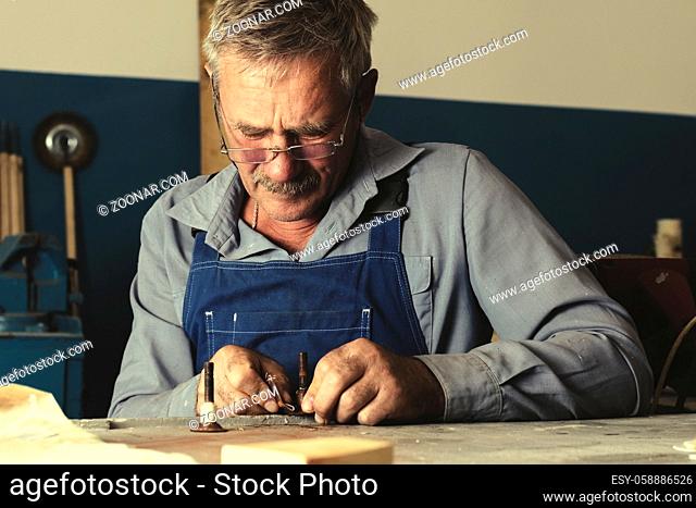 An old master with glasses works in the workshop. A white elderly man in work clothes sits at a workbench and carefully examines a metal part