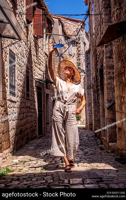 Beautiful blonde young female traveler wearing straw sun hat, taking selfie while sightseeing and enjoying summer vacation in an old traditional costal town at...