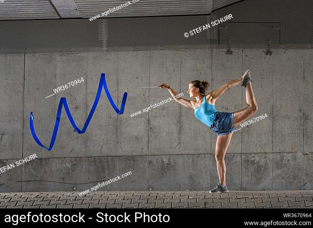 Young woman spinning ribbon standing on one leg against concrete wall under bridge