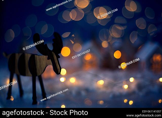 Christmas wooden reindeer over small glowing toy houses village garland over bokeh lights background with copy space for text