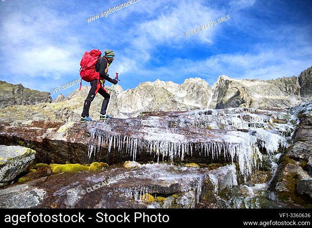 Hiker crossing a frozen creek along Sentiero Roma, Valle del Ferro, Val Masino, Valtellina, Sondrio province, Lombardy, Italy