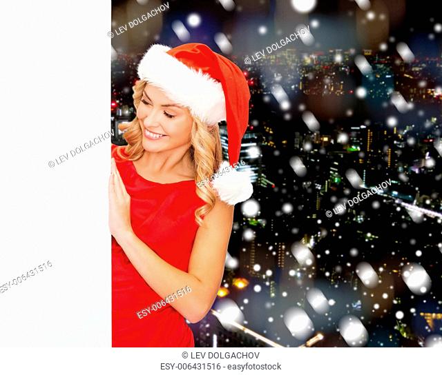 winter holidays, christmas, advertising and people concept - smiling young woman in santa helper hat with white blank billboard over snowy night city background