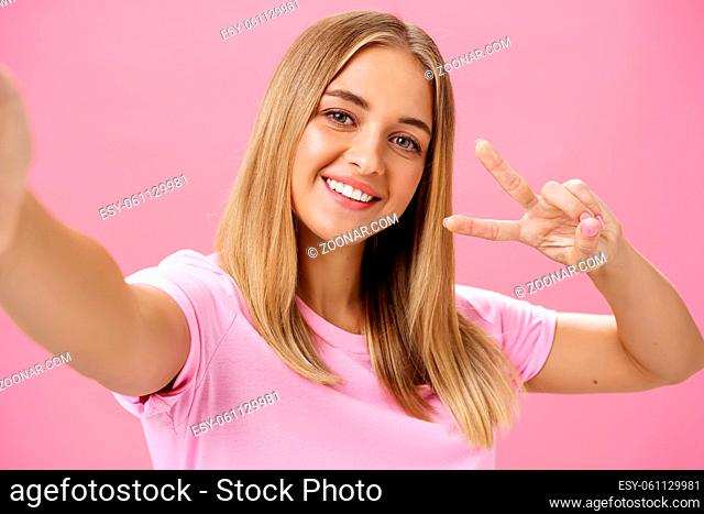 Charming friendly-looking kind young female student with fair hair and chubby face pulling hand towards to hold camera showing peace gesture and smiling broadly...