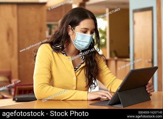 Young ethnic woman wearing yellow sweater with black and white striped blouse and face mask, sitting at bar in kitchen of downtown loft with iPad