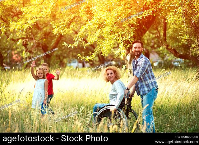 Beautiful Woman In Wheelchair With Her Husband Holding The Push Handles Look Back And Smiles At Somebody. Two Charming Kids Are Waving