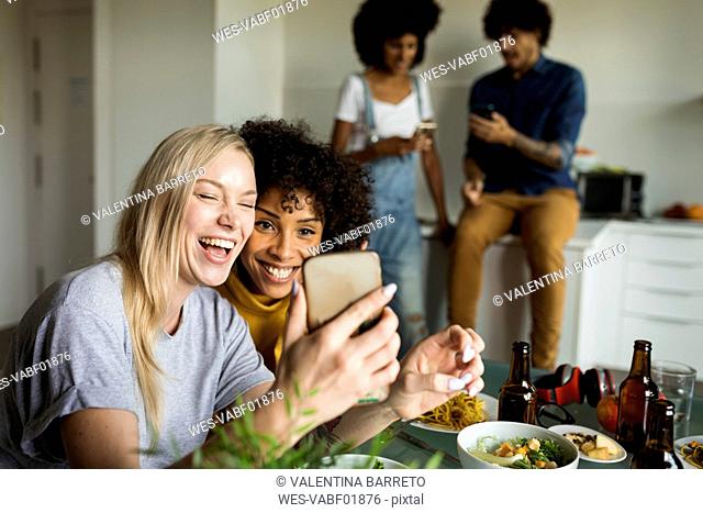 Happy girlfriends with cell phone sitting at dining table