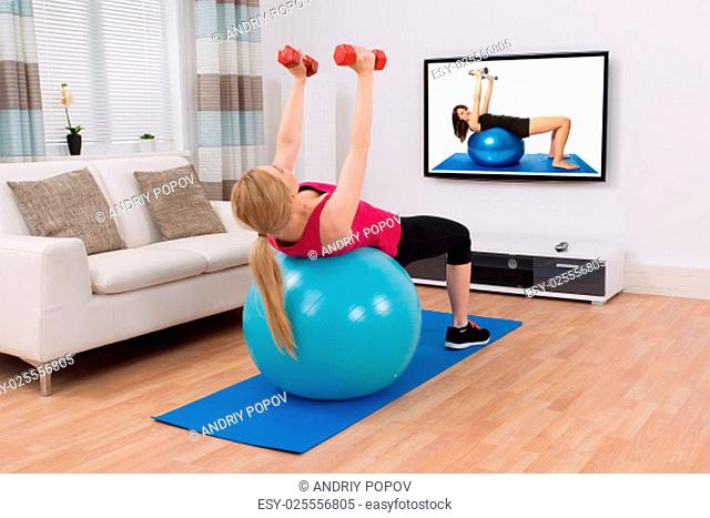 Young Woman Exercising With Fitness Ball And Dumbbell In Front Of Television