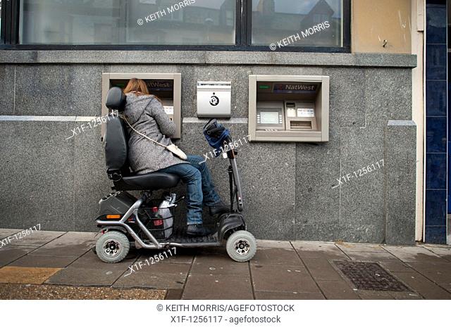 a woman in a powered wheelchair scooter using an ATM cash machine, UK