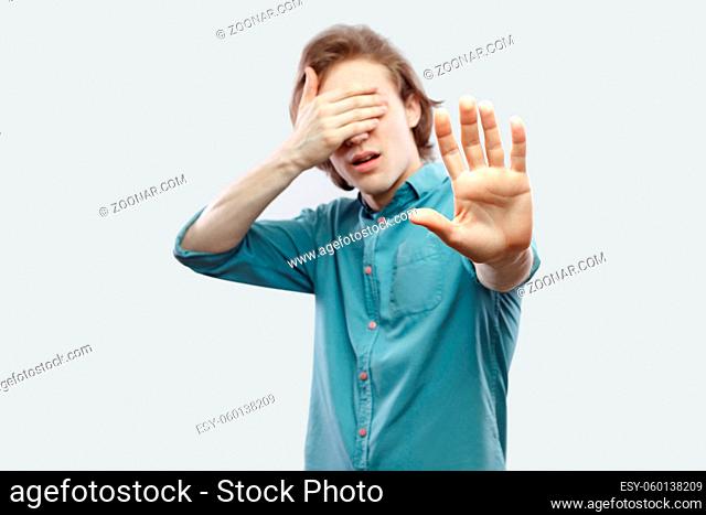Stop, I don't to see this. Portrait of handsome long haired blonde young man in blue casual shirt standing with block hand and covering his eyes