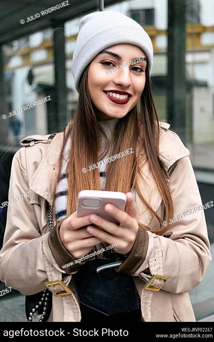 Mujer sonriente con teléfono inteligente apoyado en la pared de cristal