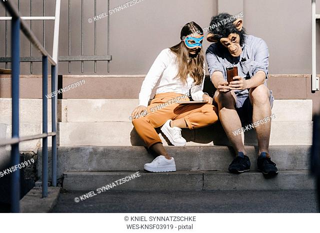 Couple in disguise sitting on stairs, using smartphone