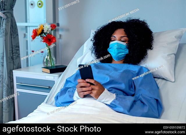 African american female patient lying in hospital bed wearing face mask using smartphone