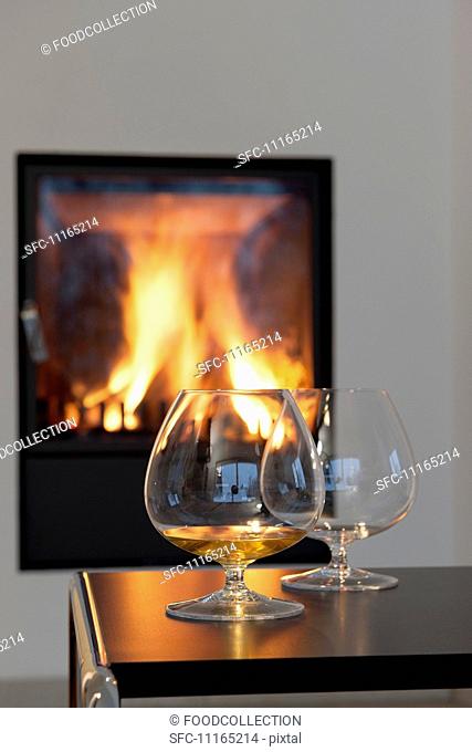 Two cognac snifters on a table in front of a roaring fire in a fireplace
