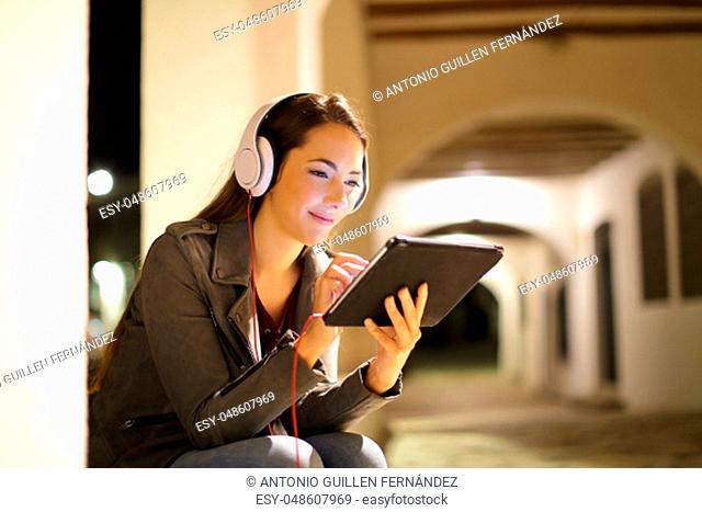 Relaxed woman wearing headphones watching media in a tablet in the night in the street