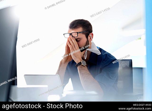 Young entrepreneur with protective face mask sitting with head in hands at office