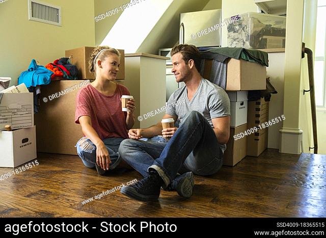 Young couple with coffee sitting on floor with boxes while moving in to new home