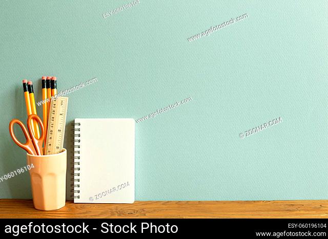 White notebook and pencils, ruler and scissors in holder on wooden desk. blue wall background. workspace