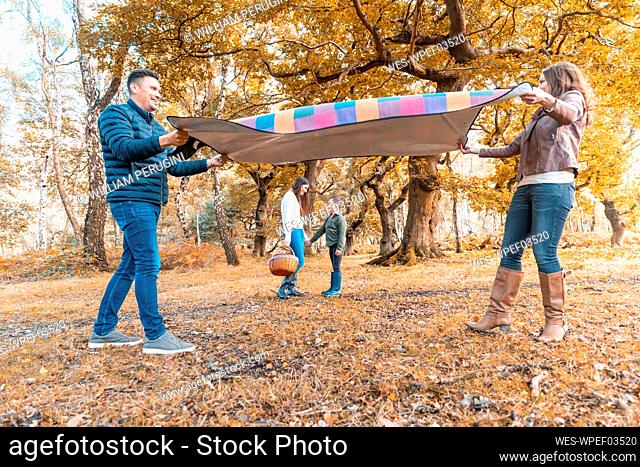 Parents laying down picnic blanket while children standing behind in park during autumn