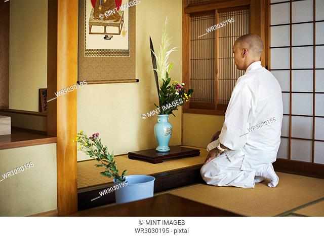Side view of Buddhist monk with shaved head wearing white robe kneeling in front of vase with flowers