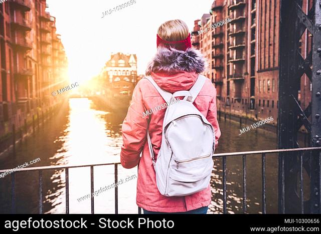 Rear view of adult woman tourist with white backpack enjoying sunset on the bridge in Speicherstadt historical warehouse district