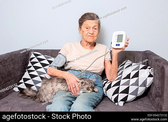 The topic very old person and monitor health. senior Caucasian woman, 90 years old, with wrinkles and gray hair, sits home on a sofa with a pet cat and uses a...