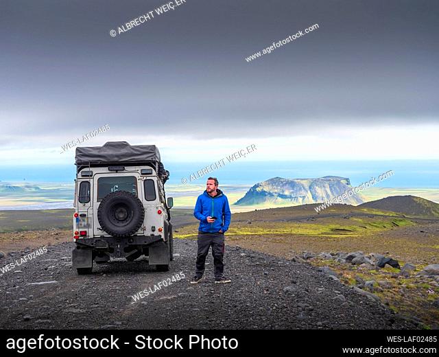Male tourist looking away while standing by off-road vehicle on road against cloudy sky