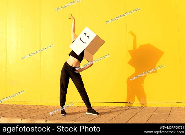Woman wearing box with smiley face dancing in front of of yellow wall