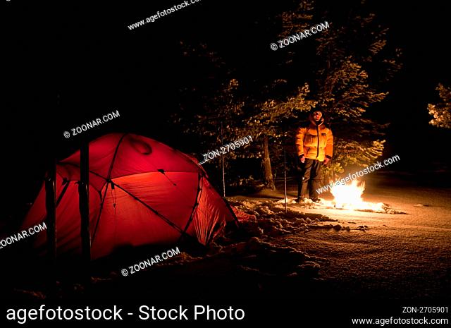 Mann am Lagerfeuer neben einem erleuchteten Zelt im Schnee, Femundsmarka, Hedmark Fylke, Norwegen, Dezember 2010