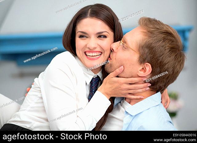 Close up of happy couple at home. Gorgeous lady smiling wide while being kissed by man. Man wearing clear glasses kissing his beautiful wife on cheek gently...