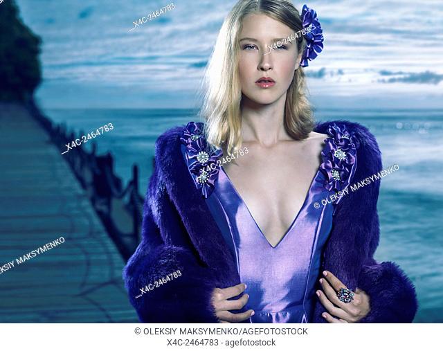 Beauty portrait of a young beautiful blond woman wearing a blue evening dress and a fur jacket outdoors at waterfront