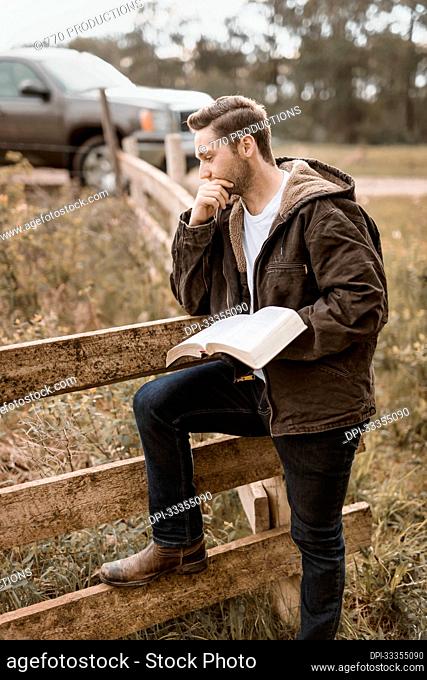 A young man stands holding his open Bible beside a rail fence and truck park on a road in the countryside; Alberta, Canada