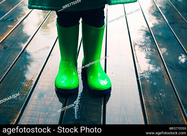 Closeup of legs of kid standing on a porch wearing green wellies and raincoat. Rain boots in bright green color