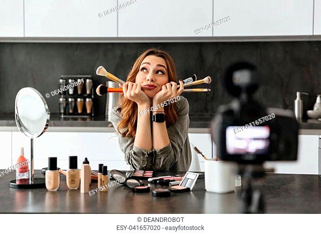 Thoughtful young girl recording her video blog episode about new cosmetic products while sitting at the kitchen table at home and applying make-up