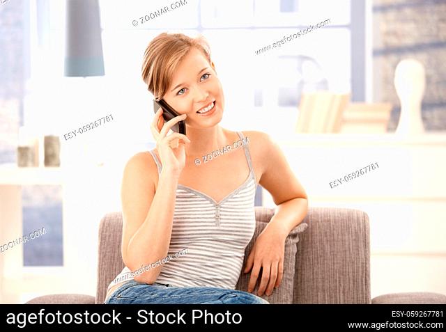 Young woman sitting on sofa at home, talking on mobile phone, smiling