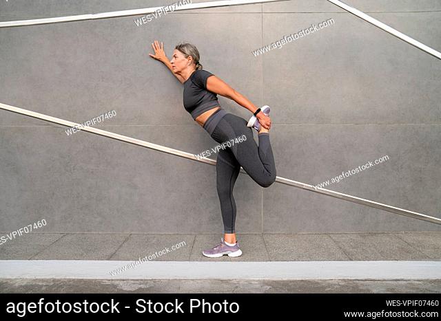 Woman stretching leg on footpath by gray wall