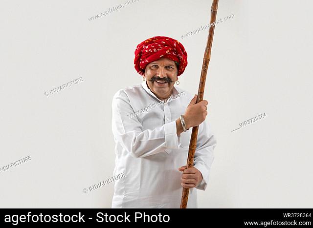 A RURAL MAN HAPPILY STANDS IN FRONT OF CAMERA WITH A LONG STICK IN HAND