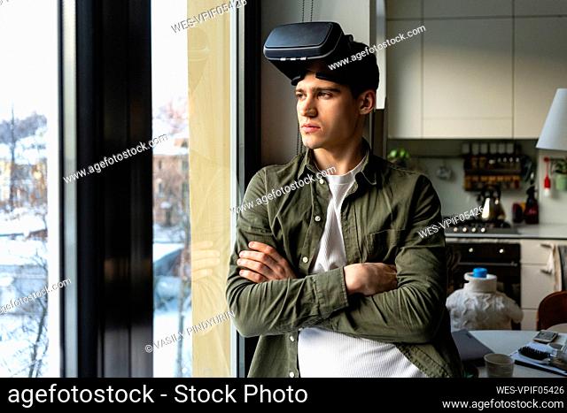 Young man wearing virtual reality simulator standing with arms crossed looking through window