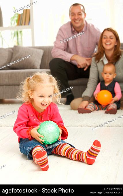 Smiling little girl holding ball at home with laughing family in background