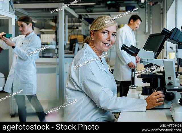 Smiling scientist sitting by microscope with coworkers working in background at laboratory