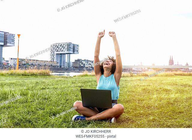 Germany, Cologne, cheering woman sitting on a meadow using laptop