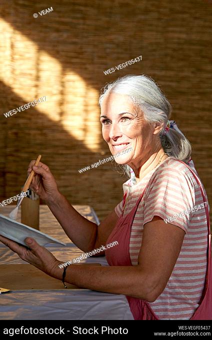 Smiling woman painting clay plate sitting at table