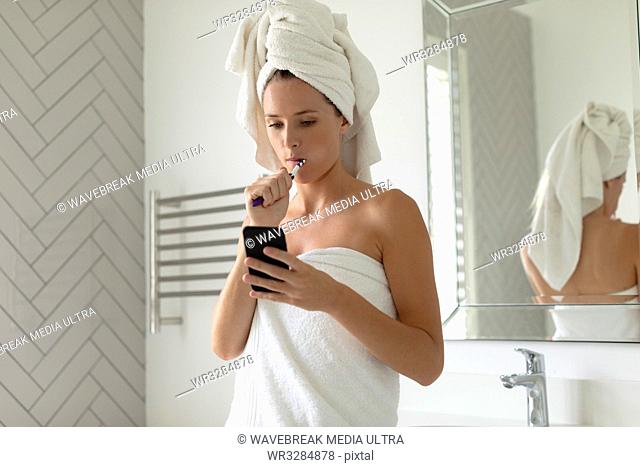 Beautiful woman using her phone while brushing her teeth in bathroom