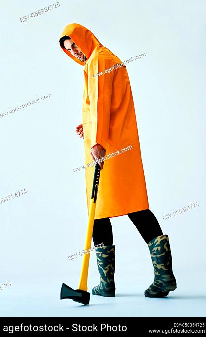 Full length portrait of young spooky man in bright raincoat holding big axe on white background