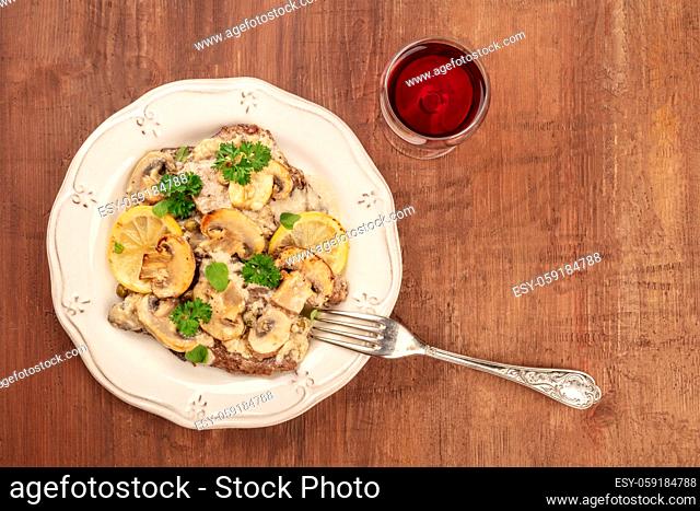 Overhead photo of scallopine di vitello, veal scallopini, with mushroom sauce, lemons, and parsley, shot from above on dark rustic texture with a glass of red...