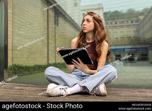 Thoughtful woman holding digital tablet while sitting in front of glass wall