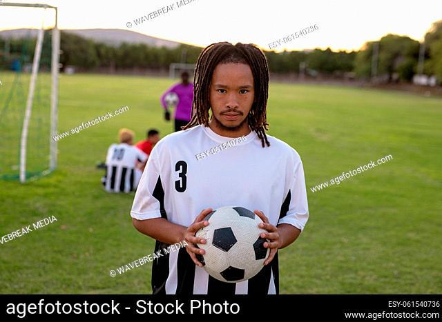 Portrait of serious biracial male player in white uniform holding soccer ball standing at playground