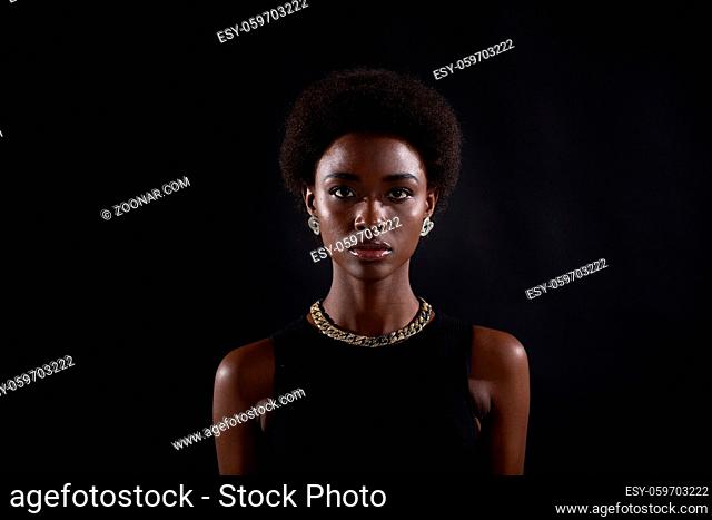 Portrait of young serious stylish black woman. Beautiful curly dark haired african american girl wear black dress with gold jewelry and looking at camera