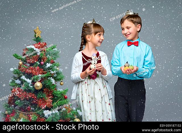 Beautiful little boy and girl standing near christmas tree, holding party dessert. Over snow backgroud